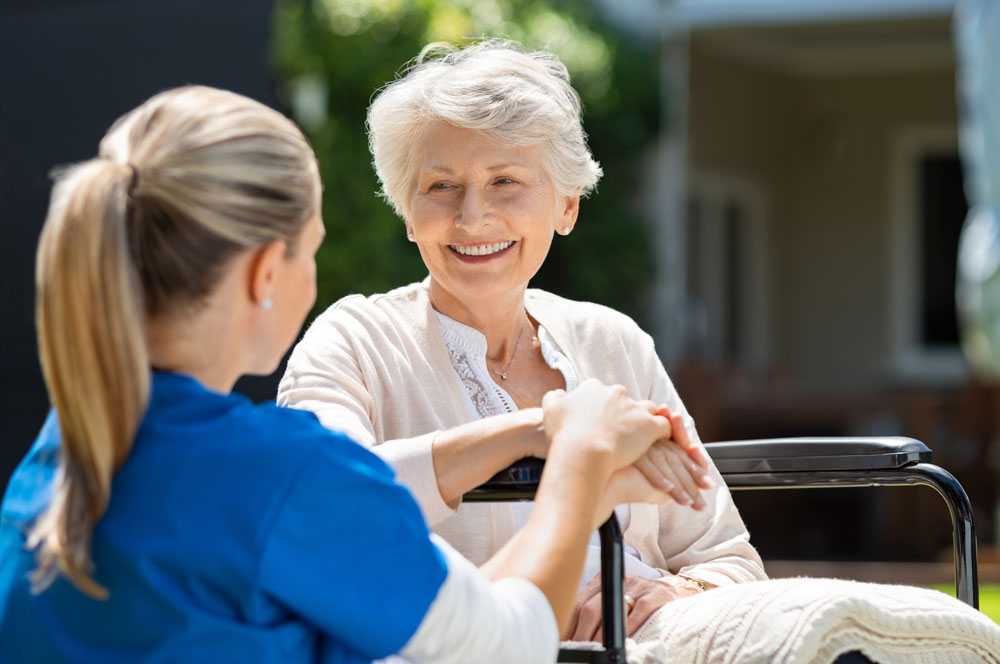 Junge Krankenpflegerin sitzt im Freien auf einer Bank und spricht mit einer lächelnden älteren Frau